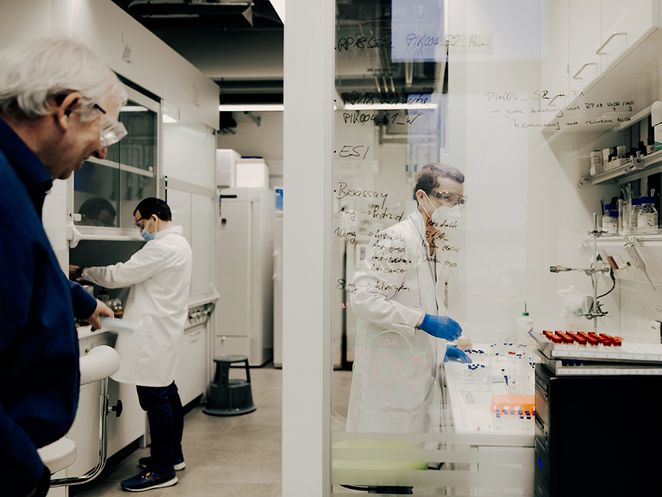 Zwei Wissenschaftler arbeiten in weißen Kitteln, mit Schutzbrille und Handschuhen in einem hellen Labor, Ludger Wessjohann im Vordergrund.