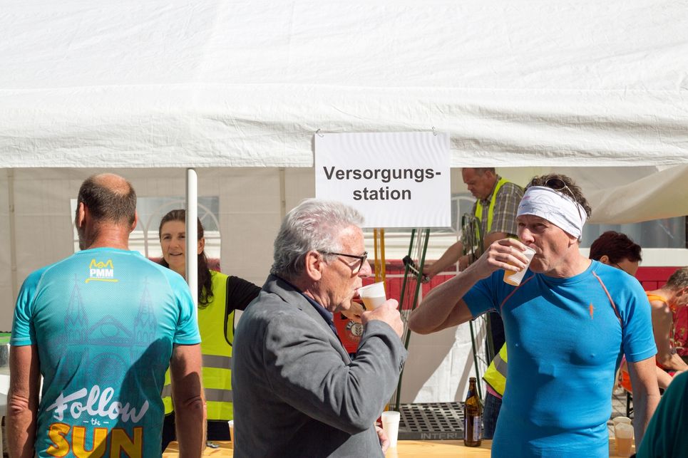 Personen trinken Bier an der einem weißen Pavillon mit der Aufschrift: Versorgungsstation.