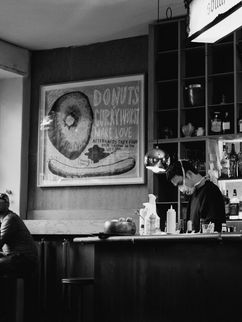 Bar, in der zwei Personen am Fenster sitzen, ein Barkeeper mit Mund-Nasen-Schutz.