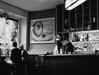 Bar, in der zwei Personen am Fenster sitzen, ein Barkeeper mit Mund-Nasen-Schutz.