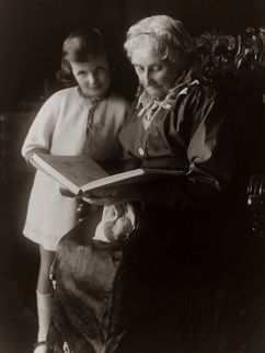 Historische schwarzweiß Fotografie einer älteren Dame, die einem jungen Mädchen vorliest, das neben ihr steht.