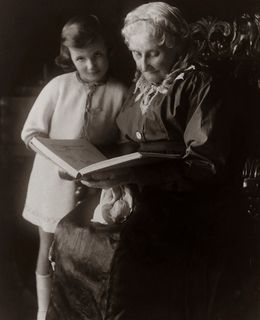 Historische schwarzweiß Fotografie einer älteren Dame, die einem jungen Mädchen vorliest, das neben ihr steht.