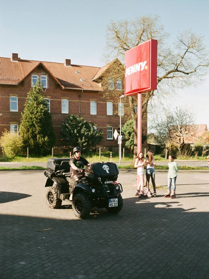 Ein schwarz gekleideter Mann in einem Squat mit aufgemaltem Totenkopf und drei Mädchen auf einem Penny-Parkplatz.