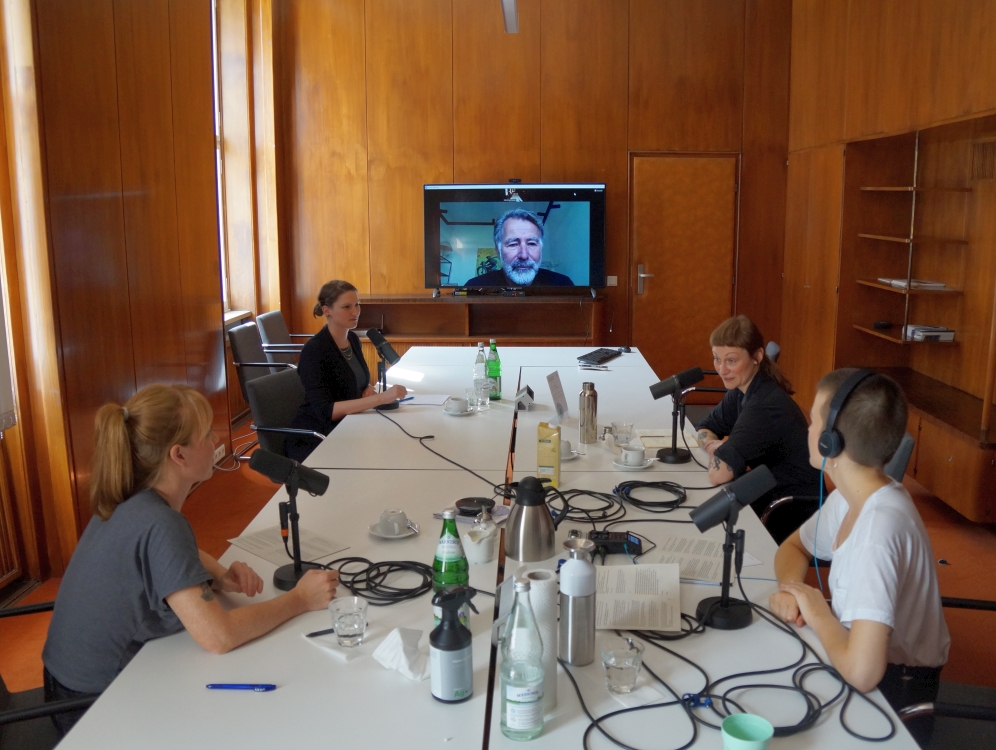 Vier Personen in einem Konferenzraum, eine weitere Person ist per Video zugeschaltet. Auf dem Tisch vor ihnen Mikrophone, Kabel, Notizzettel, Getränke.
