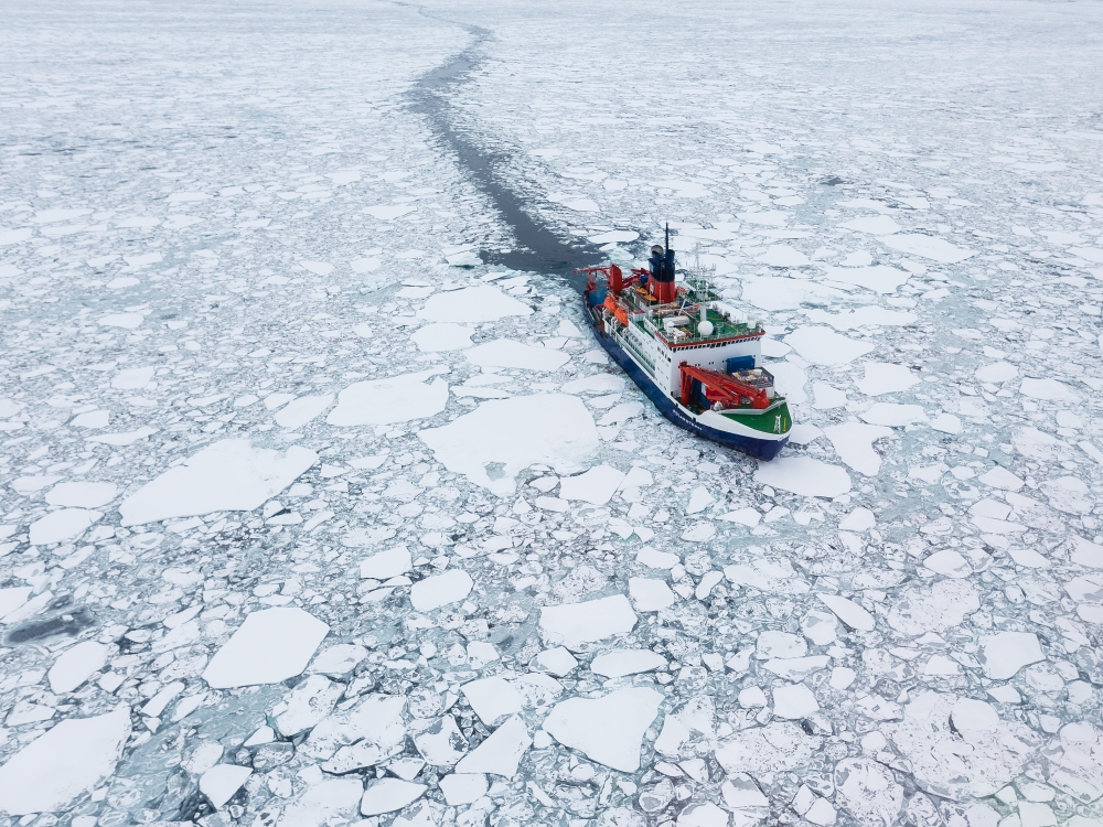 Ein großes Schiff, ein Eisbrecher, fährt durch Eisschollen und hinterlässt eine dunkle Fahrrinne.