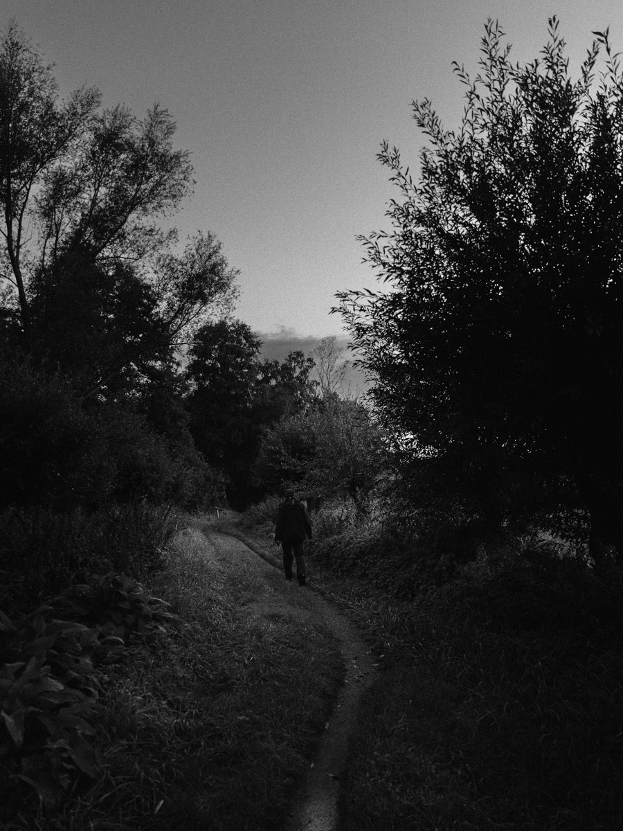 Ein Mann geht einen dicht bewaldeten Feldweg entlang. Das Bild ist in schwarz-weiß.