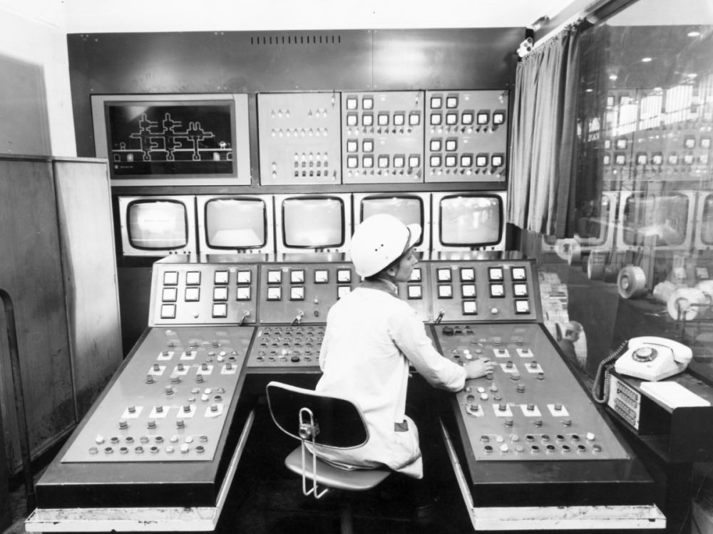 In einem Kontrollraum sitzt eine Person vor einem großen Bedienpult mit zahlreichen Schaltern, Anzeigen und Bildschirmen. Die Schwarzweißaufnahme zeigt einen technischen Schaltraum in einem Industriebetrieb.
