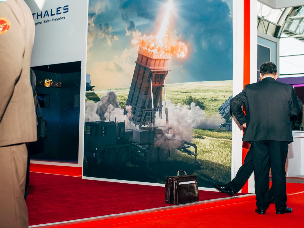 Zwei Herren in Anzügen, einer in Uniform for einem Plakat einer Raketenabwehr-Maschine.zu sehen und einer in Uniform, der ebenfalls nur halb zu sehen ist, von