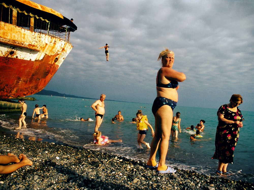 Badende an einem steinigen Strand, ein altes Schiff wird als Sprungturm genutzt. Trotz bewölktem Himmel scheint die Sonne auf das türkisblaue Wasser.