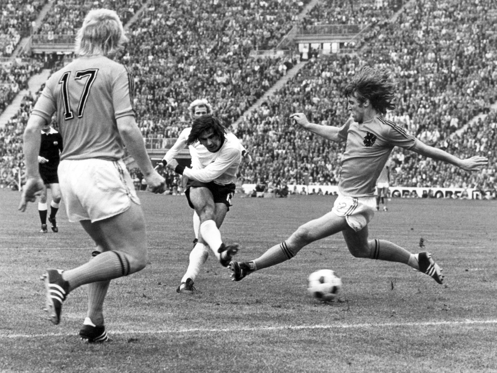 Schwarz-weiß Fotografie zeigt drei Fußballer beim Fußballspielen vor voll besetzten Rängen.