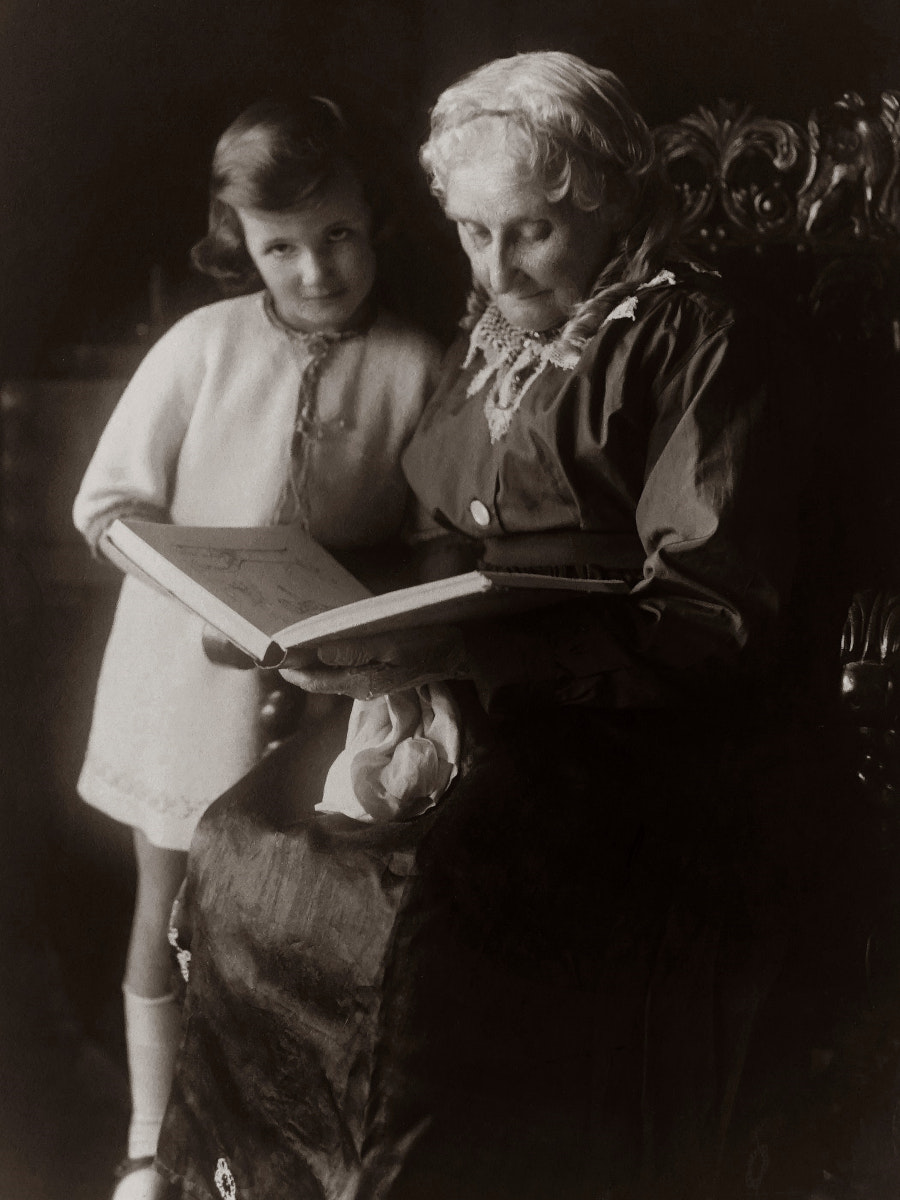 Historische schwarzweiß Fotografie einer älteren Dame, die einem jungen Mädchen vorliest, das neben ihr steht.