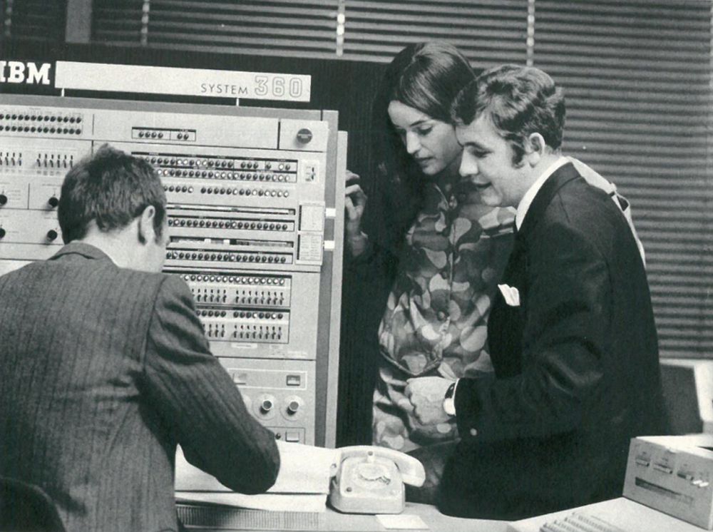 Junges Pärchen, Frau und Mann vor einem Schreibtisch, an dem ein weiterer Mann sitzt. Neben ihnen ein Kasten mit vielen Knöpfen. Alles im Stil der 1960er, 1970er Jahre.