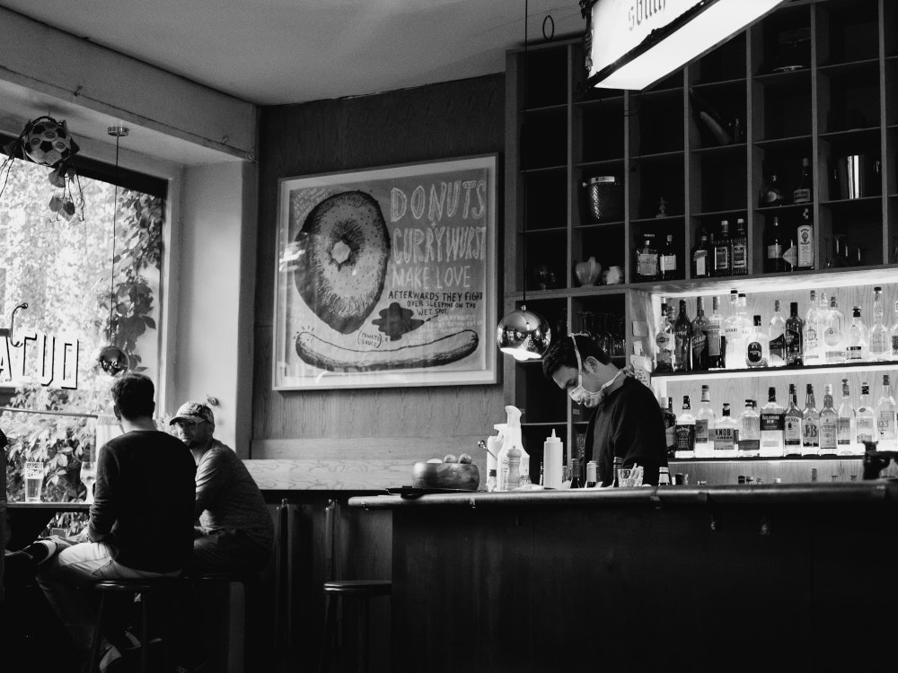 Bar, in der zwei Personen am Fenster sitzen, ein Barkeeper mit Mund-Nasen-Schutz.